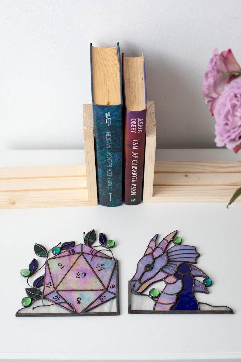 May include: A wooden bookend holding two books with blue and purple covers. Below, two stained glass bookends: a D20 die and a dragon head, both with purple, green, and blue accents. The die has numbers and the dragon has a detailed design.