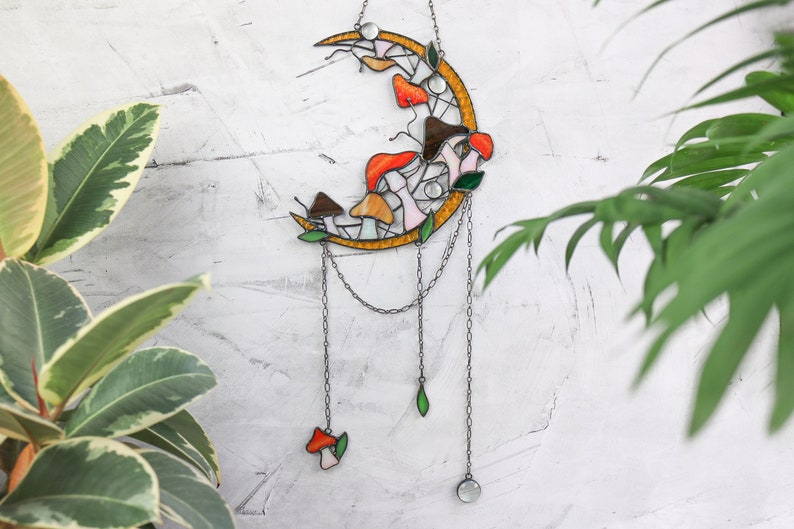 Puede incluir: Una luna creciente de vidrio tintado con una cadena colgando de ella. La luna est&aacute; decorada con setas coloridas en tonos naranja, amarillo y blanco. La cadena tiene una peque&ntilde;a hoja verde y un cristal transparente en el extremo.