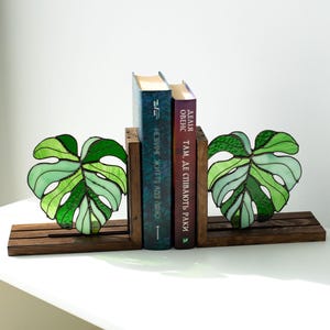 K&ouml;nnte beinhalten: Buchst&uuml;tzen in Form von Monstera-Bl&auml;ttern aus gr&uuml;nem Glas, auf einem dunklen Holzsockel. Zwei B&uuml;cher stehen zwischen den Buchst&uuml;tzen. Ein dekoratives Element f&uuml;r jedes B&uuml;cherregal.
