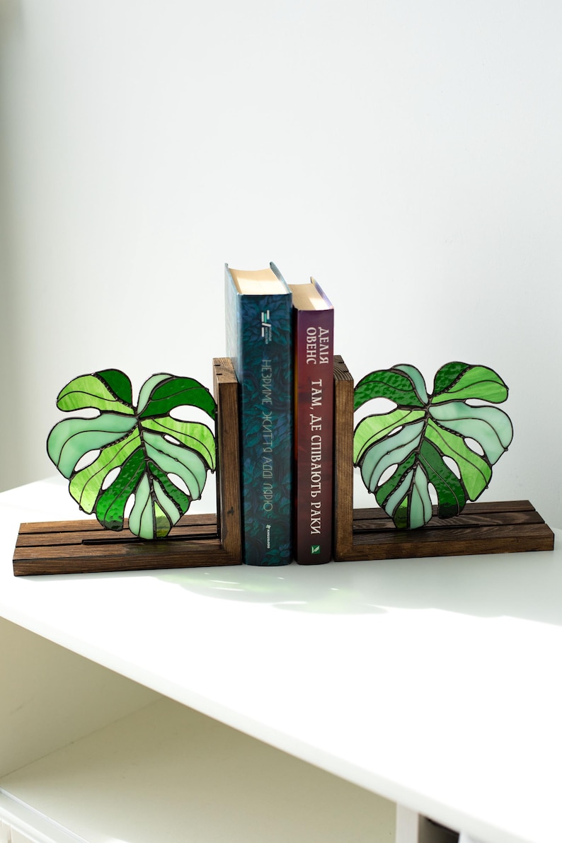 K&ouml;nnte beinhalten: Buchst&uuml;tzen in Form von Monstera-Bl&auml;ttern aus gr&uuml;nem Glas, auf einem dunklen Holzsockel. Zwei B&uuml;cher stehen zwischen den Buchst&uuml;tzen. Ein dekoratives Element f&uuml;r jedes B&uuml;cherregal.