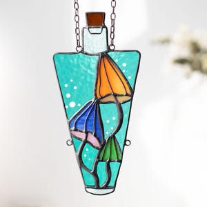 May include: A stained glass suncatcher shaped like a bottle with a turquoise blue background and three colorful mushrooms inside. The mushrooms are orange, blue, and green.