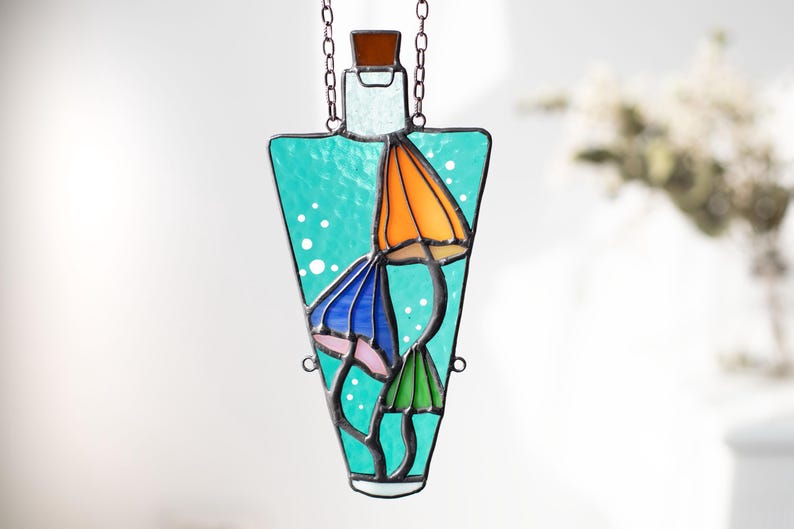May include: A stained glass suncatcher shaped like a bottle with a turquoise blue background and three colorful mushrooms inside. The mushrooms are orange, blue, and green.