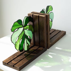 K&ouml;nnte beinhalten: Dunkel gebeizte Holz-Buchst&uuml;tzen mit einem Buntglas-Monstera-Blatt-Design in Gr&uuml;nt&ouml;nen. Die Buchst&uuml;tzen haben eine rechteckige Basis und vertikale St&uuml;tzen und schaffen so einen dekorativen und funktionalen Artikel.