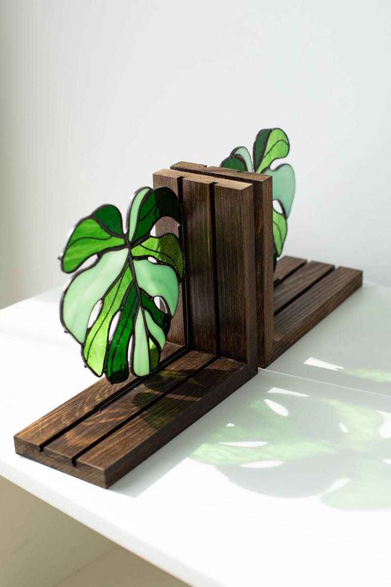 K&ouml;nnte beinhalten: Dunkel gebeizte Holz-Buchst&uuml;tzen mit einem Buntglas-Monstera-Blatt-Design in Gr&uuml;nt&ouml;nen. Die Buchst&uuml;tzen haben eine rechteckige Basis und vertikale St&uuml;tzen und schaffen so einen dekorativen und funktionalen Artikel.