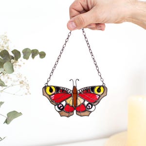 May include: A stained glass butterfly with red, yellow, and brown details, suspended by a chain. The butterfly has a symmetrical design with intricate patterns. The wings are primarily red with yellow and brown accents.