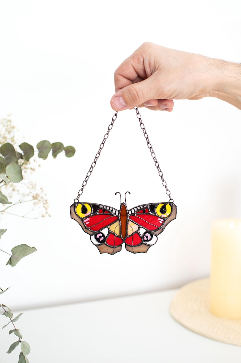 May include: A stained glass butterfly with red, yellow, and brown details, suspended by a chain. The butterfly has a symmetrical design with intricate patterns. The wings are primarily red with yellow and brown accents.