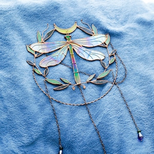 May include: A stained glass dragonfly suncatcher with iridescent wings, a crescent moon, and leafy branches. The suncatcher hangs from three chains with blue and green beads.