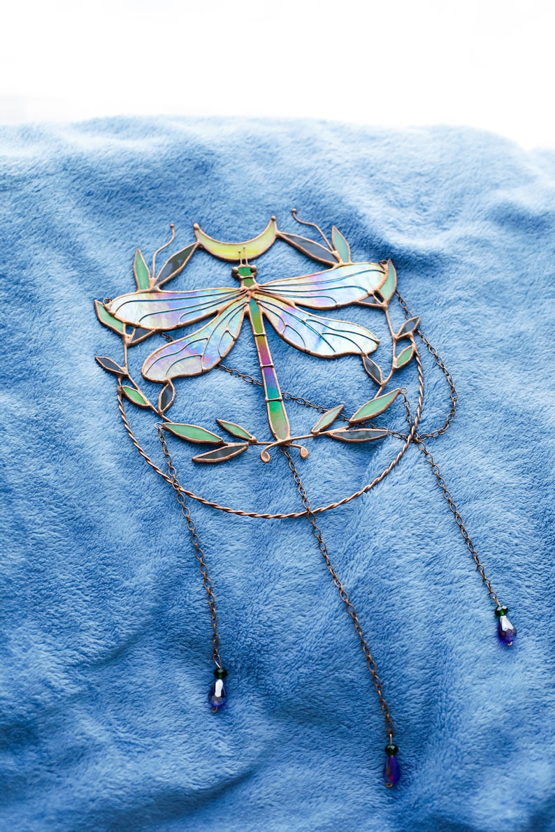 May include: A stained glass dragonfly suncatcher with iridescent wings, a crescent moon, and leafy branches. The suncatcher hangs from three chains with blue and green beads.
