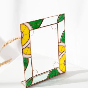 May include: A square stained glass frame with a copper-colored border. The frame features a design of yellow, green, and white segments, resembling citrus fruit slices and leaves. The frame is standing upright on a white surface.