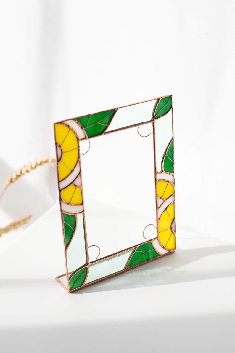 May include: A square stained glass frame with a copper-colored border. The frame features a design of yellow, green, and white segments, resembling citrus fruit slices and leaves. The frame is standing upright on a white surface.