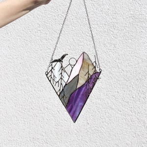 May include: A stained glass window hanging from a chain. The window is shaped like a diamond and depicts a purple, pink, and gray mountain scene with a bird flying overhead.