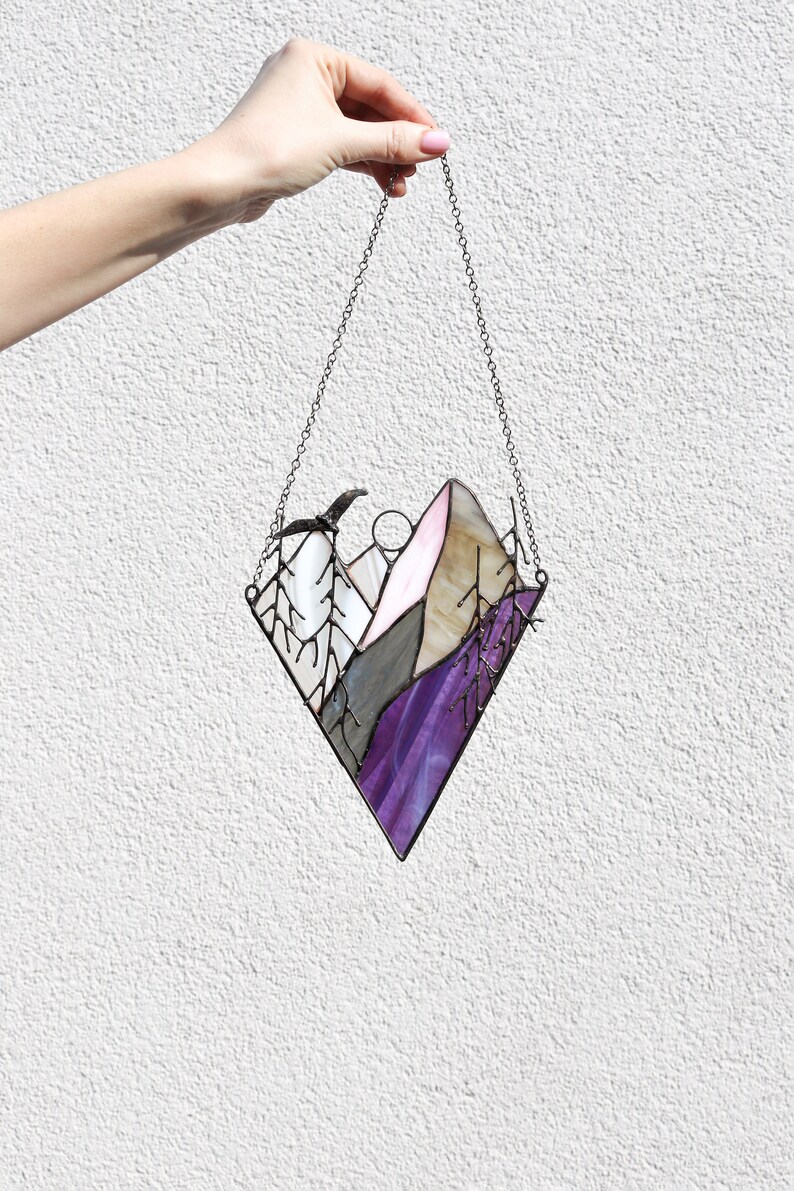 May include: A stained glass window hanging from a chain. The window is shaped like a diamond and depicts a purple, pink, and gray mountain scene with a bird flying overhead.