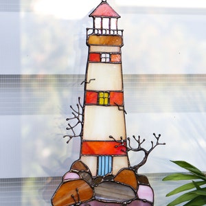 May include: A stained glass lighthouse hanging ornament with red, orange, yellow, and blue accents. The lighthouse is surrounded by brown and pink rocks.