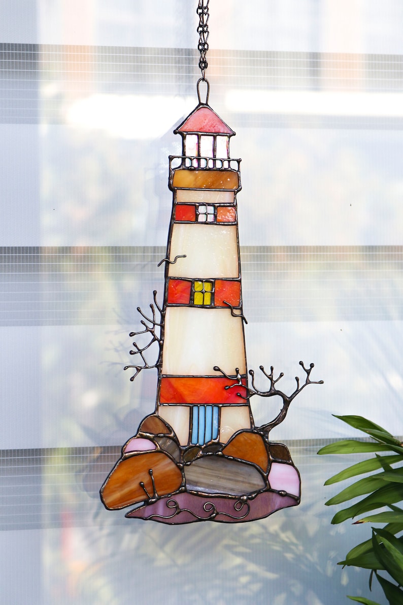 May include: A stained glass lighthouse hanging ornament with red, orange, yellow, and blue accents. The lighthouse is surrounded by brown and pink rocks.