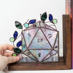 May include: A stained glass D20 die with iridescent glass, featuring numbers and a decorative vine with green and blue leaves. The die is set on a wooden base, perfect for tabletop gaming enthusiasts.
