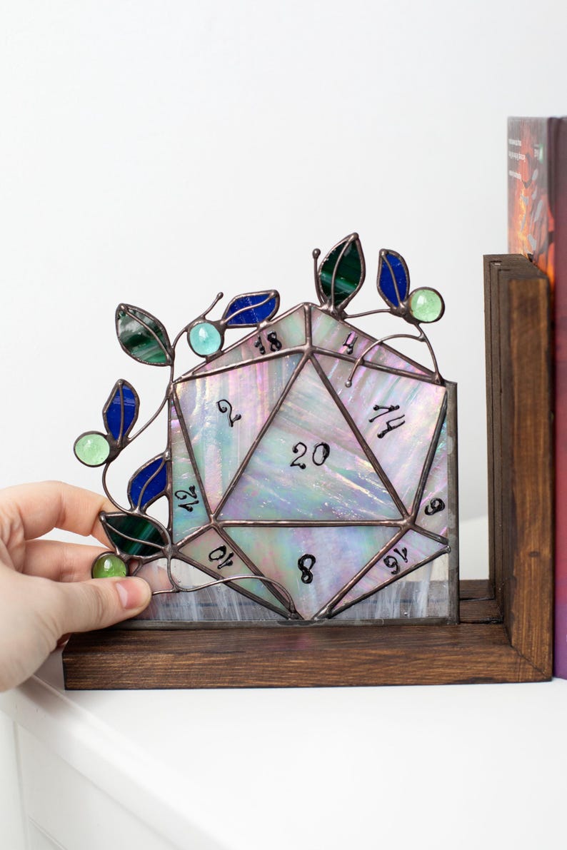 May include: A stained glass D20 die with iridescent glass, featuring numbers and a decorative vine with green and blue leaves. The die is set on a wooden base, perfect for tabletop gaming enthusiasts.
