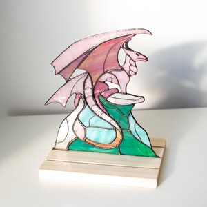 Op de afbeelding: Een gebrandschilderde drakenbeeldhouwwerk in tinten roze, rood, groen en blauw. De draak zit bovenop een kleurrijke bergsc&egrave;ne, gemonteerd op een houten voet. De vleugels zijn gespreid en de draak kijkt naar rechts.