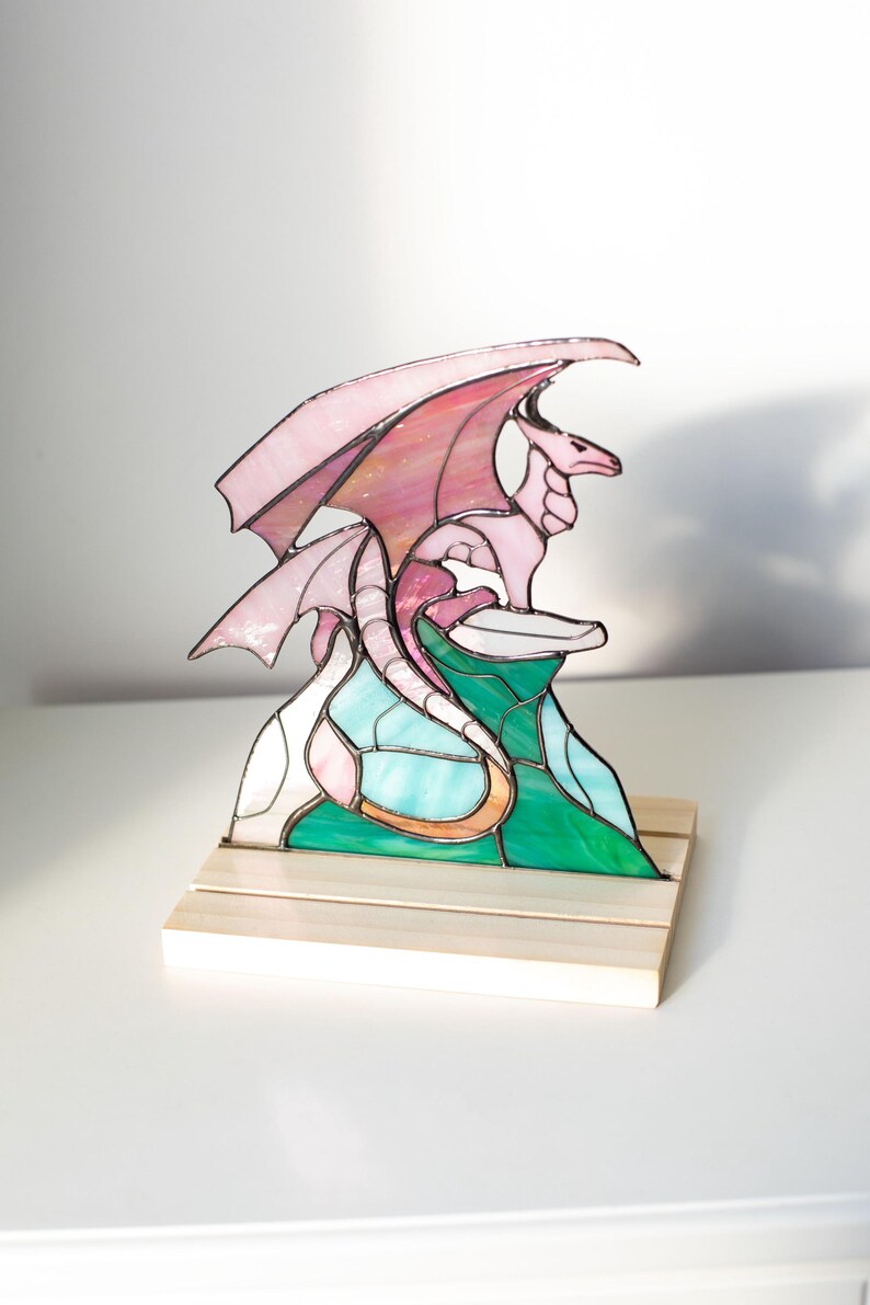 Op de afbeelding: Een gebrandschilderde drakenbeeldhouwwerk in tinten roze, rood, groen en blauw. De draak zit bovenop een kleurrijke bergsc&egrave;ne, gemonteerd op een houten voet. De vleugels zijn gespreid en de draak kijkt naar rechts.