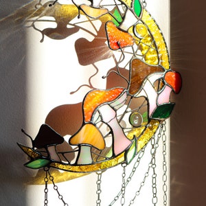 Puede incluir: Un atrapasol de cristal de colores en forma de media luna con un dise&ntilde;o de setas y hojas. El atrapasol est&aacute; colgado de una cadena y proyecta una sombra en la pared detr&aacute;s de &eacute;l.