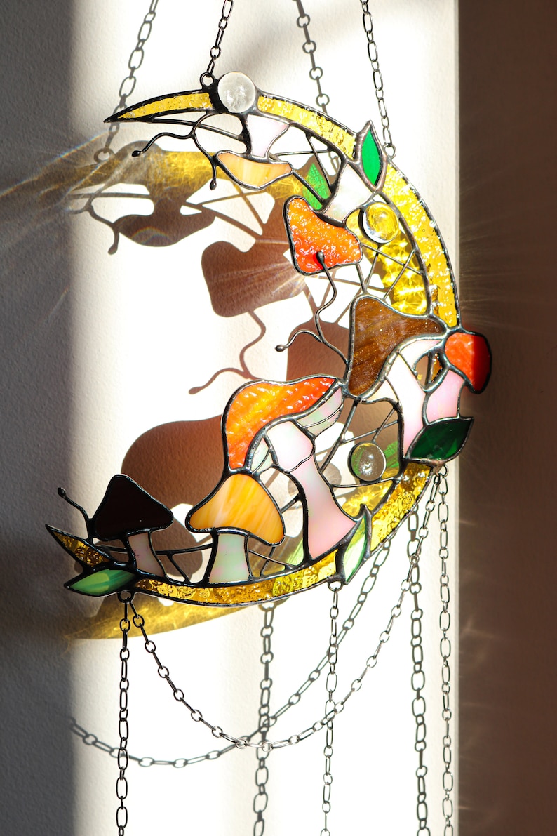 Puede incluir: Un atrapasol de cristal de colores en forma de media luna con un dise&ntilde;o de setas y hojas. El atrapasol est&aacute; colgado de una cadena y proyecta una sombra en la pared detr&aacute;s de &eacute;l.