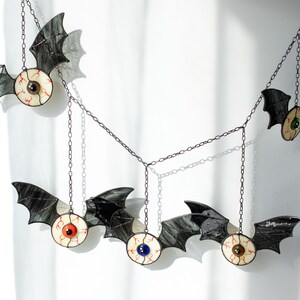 May include: A hanging mobile featuring five stained glass bats with black wings and central eyeballs. Each eyeball has a distinct iris colour including red, blue, brown, and green, with visible red veins on the white sclera. The bats are connected by thin metal chains.
