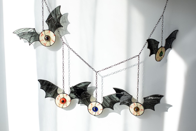 May include: A hanging mobile featuring five stained glass bats with black wings and central eyeballs. Each eyeball has a distinct iris colour including red, blue, brown, and green, with visible red veins on the white sclera. The bats are connected by thin metal chains.