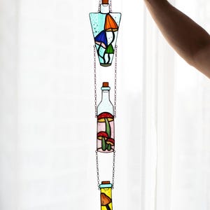 May include: Three stained glass bottles hanging from a chain. Each bottle is filled with colorful mushrooms. The bottles are in a light blue, pink, and yellow color.
