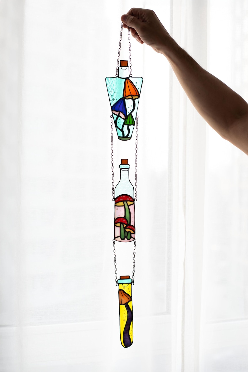 May include: Three stained glass bottles hanging from a chain. Each bottle is filled with colorful mushrooms. The bottles are in a light blue, pink, and yellow color.