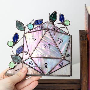 May include: Stained glass D20 die with iridescent glass and a decorative vine with blue, green, and teal accents. The die is a corner piece, with numbers visible on the facets. A unique piece for tabletop gaming enthusiasts.