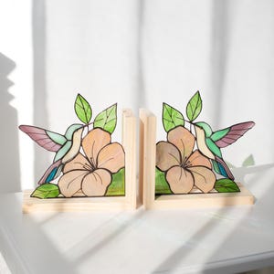 Pu&ograve; includere: Un paio di fermalibri in legno con un design a colibr&igrave;. Ogni fermalibri ha una base in legno marrone chiaro con un'illustrazione colorata di colibr&igrave; e fiori. Il colibr&igrave; ha accenti verdi, viola e blu. Il fiore &egrave; color pesca.