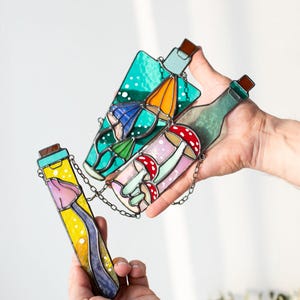 May include: Three stained glass bottles with a chain connecting them. The bottles are filled with colorful images of mushrooms, flowers, and a blue umbrella. The bottles are held in a person's hands.