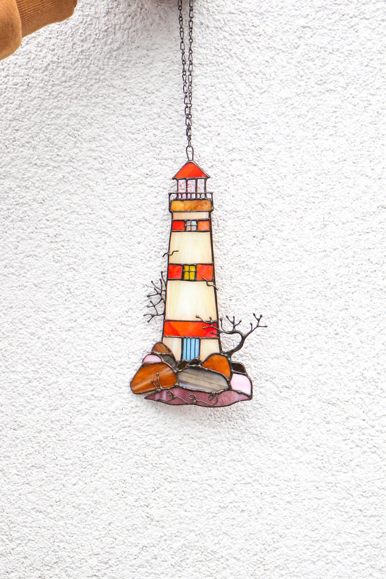 May include: A stained glass lighthouse with red, orange, and white stripes, hanging from a silver chain. The lighthouse is on a base of brown and pink rocks with a small tree on the side.