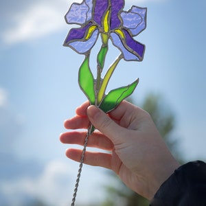May include: A stained glass iris flower with purple, blue, and green glass pieces. The flower is attached to a silver wire stem.