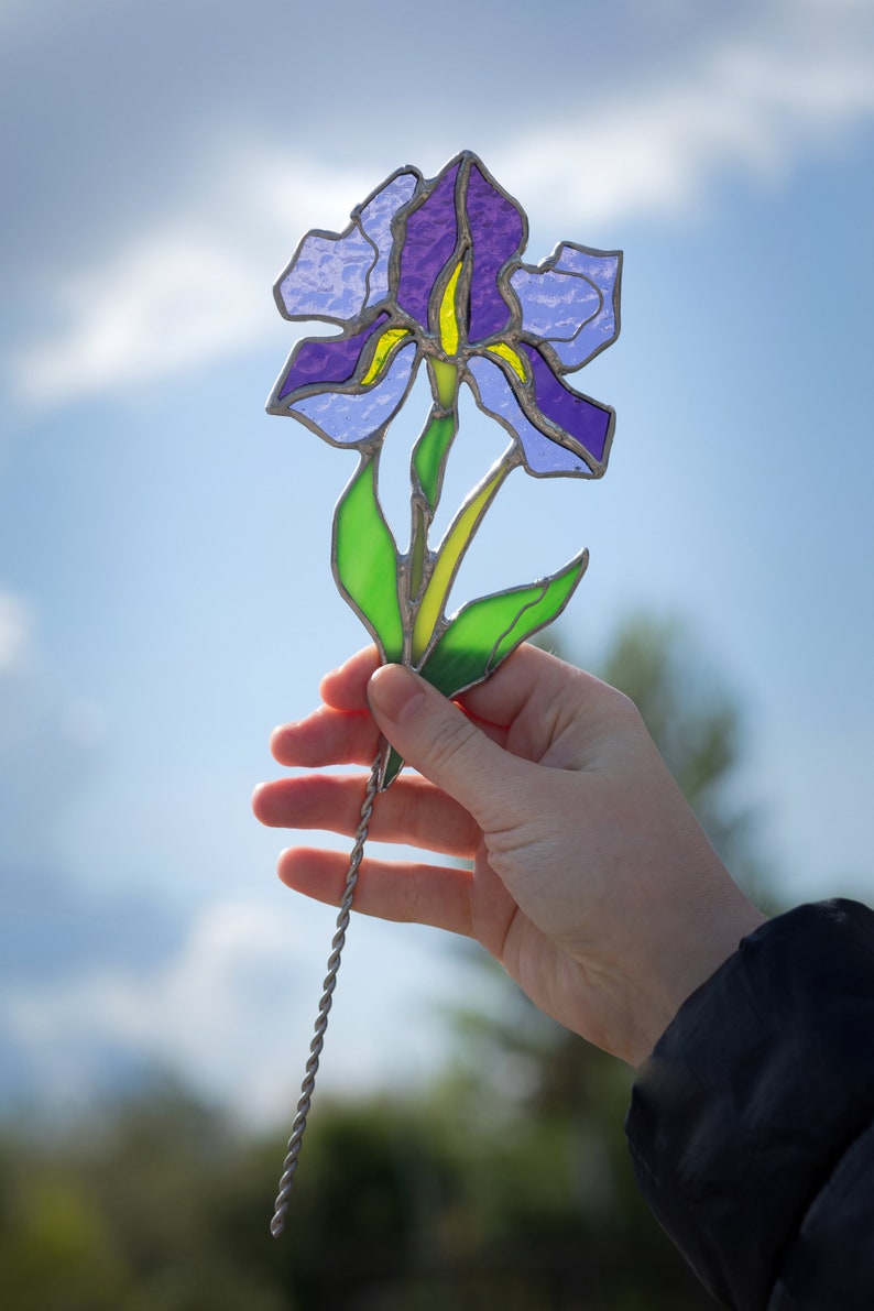 May include: A stained glass iris flower with purple, blue, and green glass pieces. The flower is attached to a silver wire stem.