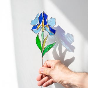 May include: A stained glass iris flower with blue, green, and yellow glass pieces. The flower is on a wire stem and is being held by a hand.