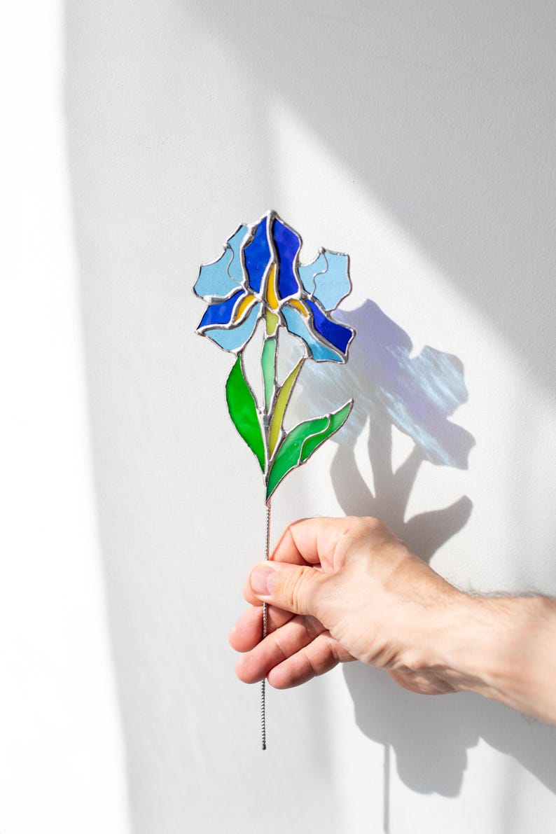 May include: A stained glass iris flower with blue, green, and yellow glass pieces. The flower is on a wire stem and is being held by a hand.