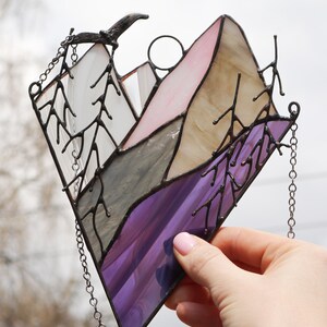 May include: A stained glass window hanging with a chain. The window depicts a purple, gray, and pink mountain scene with trees on either side.