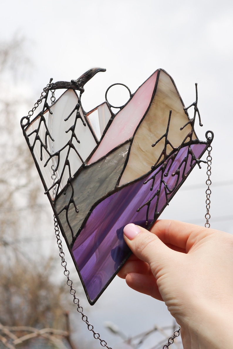 May include: A stained glass window hanging with a chain. The window depicts a purple, gray, and pink mountain scene with trees on either side.