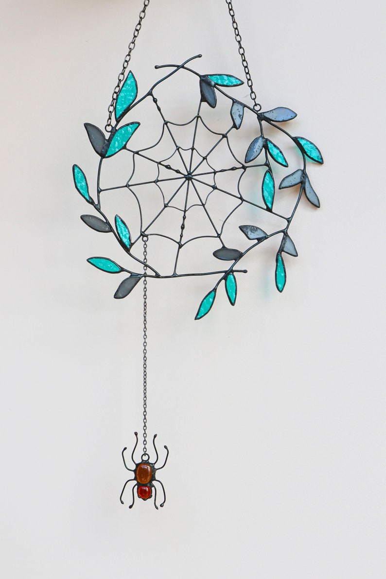 May include: A black metal spider web with teal glass leaves hanging from a chain with a small spider made of black metal and orange and red glass beads hanging from the bottom.