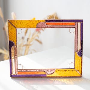 Può includere: Una cornice rettangolare in vetro colorato con un design vibrante. La cornice presenta una combinazione di pezzi di vetro giallo, arancione, viola e rosa. La cornice è larga circa 20 cm e alta 15 cm.