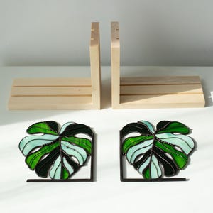 K&ouml;nnte beinhalten: Zwei Buchst&uuml;tzen-Sets. Das erste Set besteht aus hellem Holz mit rechteckiger Basis und zwei vertikalen St&uuml;tzen. Das zweite Set besteht aus zwei Buchst&uuml;tzen aus Buntglas in Monstera-Blattform mit schwarzen Metallst&uuml;tzen in Gr&uuml;n- und Wei&szlig;t&ouml;nen.