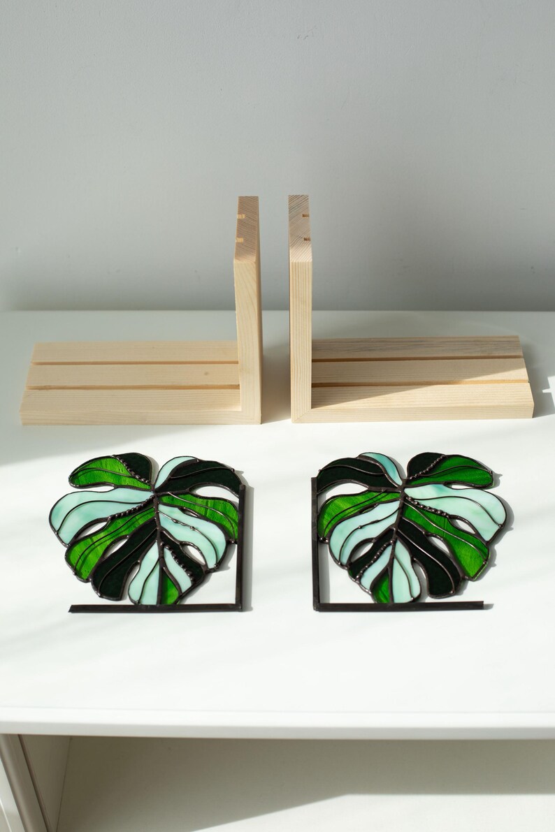 K&ouml;nnte beinhalten: Zwei Buchst&uuml;tzen-Sets. Das erste Set besteht aus hellem Holz mit rechteckiger Basis und zwei vertikalen St&uuml;tzen. Das zweite Set besteht aus zwei Buchst&uuml;tzen aus Buntglas in Monstera-Blattform mit schwarzen Metallst&uuml;tzen in Gr&uuml;n- und Wei&szlig;t&ouml;nen.
