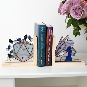 May include: Stained glass bookends featuring a D20 die and a dragon head. The bookends are set on a wooden base, with books between them. The die is iridescent with blue and green accents. The dragon is purple and blue.