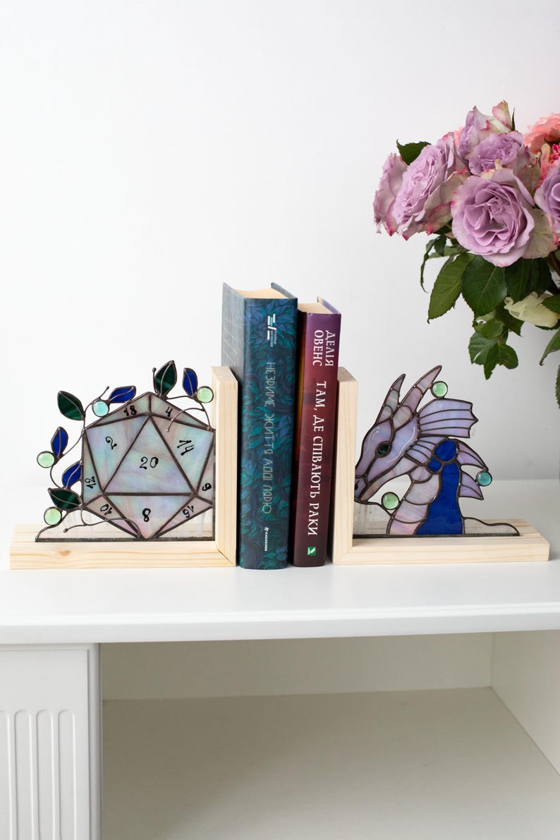 May include: Stained glass bookends featuring a D20 die and a dragon head. The bookends are set on a wooden base, with books between them. The die is iridescent with blue and green accents. The dragon is purple and blue.