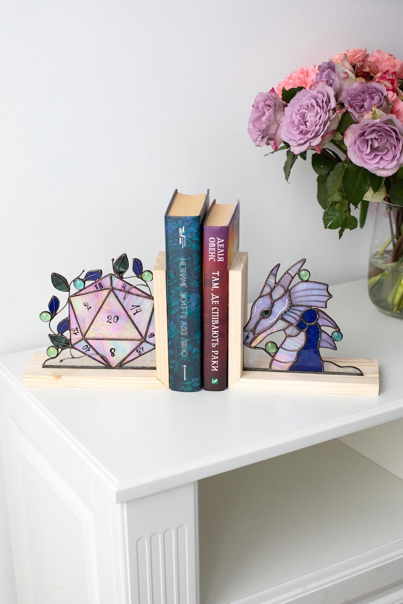 May include: Stained glass D20 dice and dragon bookends on a wooden base. The bookends are holding two books. A vase of purple roses sits on the right side of the table.