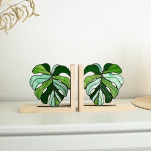 K&ouml;nnte beinhalten: Zwei Buchst&uuml;tzen aus Glas in Form von Monstera-Bl&auml;ttern. Die Bl&auml;tter sind in Gr&uuml;n- und Hellblaut&ouml;nen gehalten und mit schwarzen Konturen versehen. Sie stehen auf hellen Holzsockeln, ideal f&uuml;r B&uuml;cher.