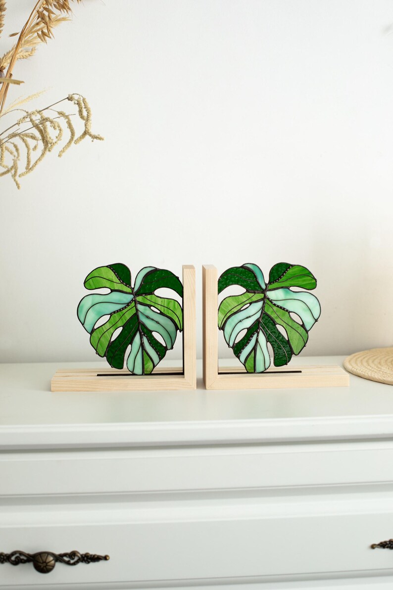 K&ouml;nnte beinhalten: Zwei Buchst&uuml;tzen aus Glas in Form von Monstera-Bl&auml;ttern. Die Bl&auml;tter sind in Gr&uuml;n- und Hellblaut&ouml;nen gehalten und mit schwarzen Konturen versehen. Sie stehen auf hellen Holzsockeln, ideal f&uuml;r B&uuml;cher.