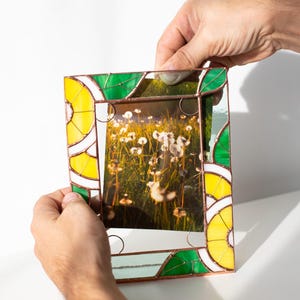 May include: A stained glass picture frame with a photo of a field of dandelions. The frame has a yellow, green, and white design. The frame is being held by two hands.