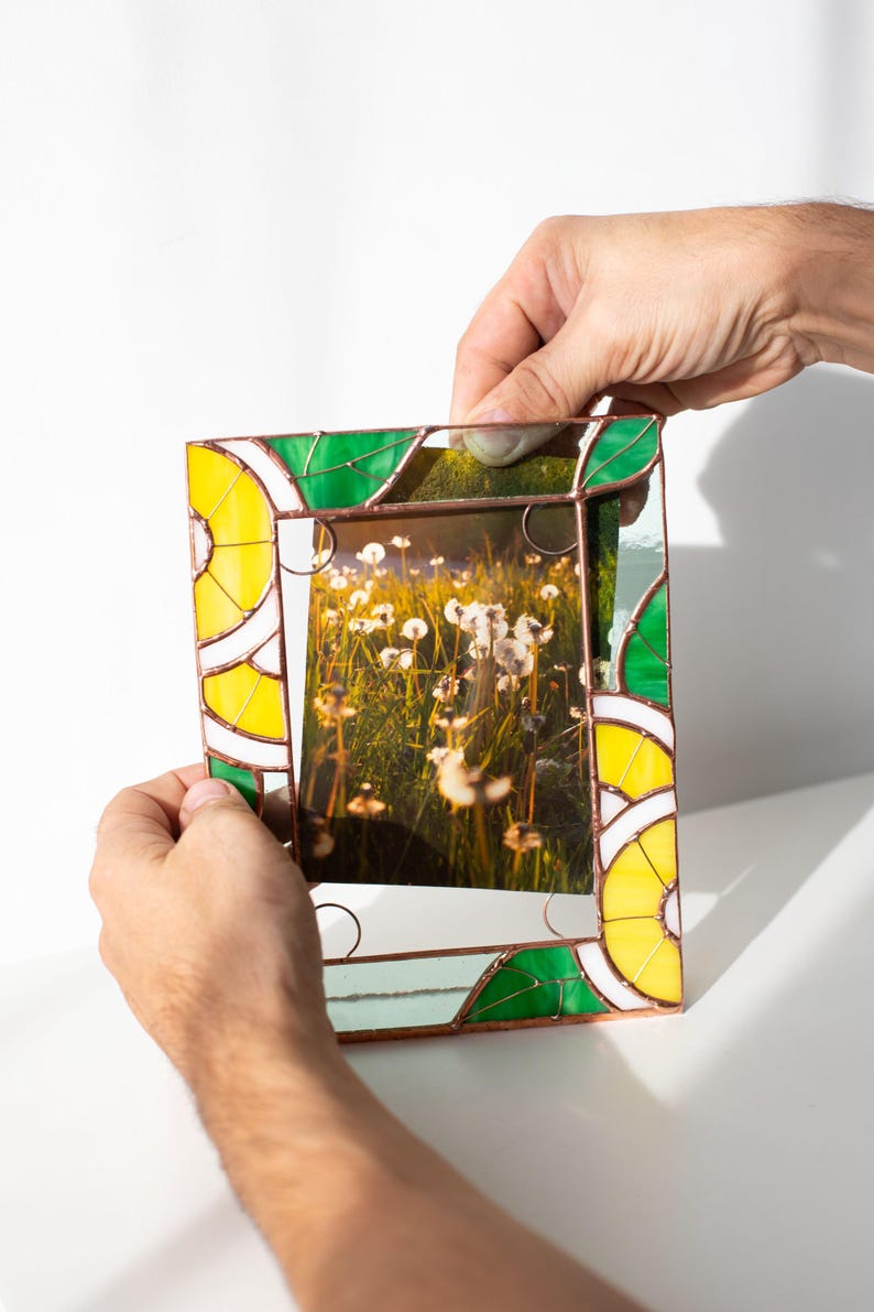 May include: A stained glass picture frame with a photo of a field of dandelions. The frame has a yellow, green, and white design. The frame is being held by two hands.