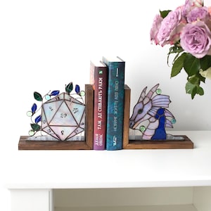May include: Stained glass bookends with a D20 die and a dragon design. The bookends are made of wood and hold two books. The die is white with black numbers, and the dragon is purple and blue. A bouquet of pink roses is in the background.